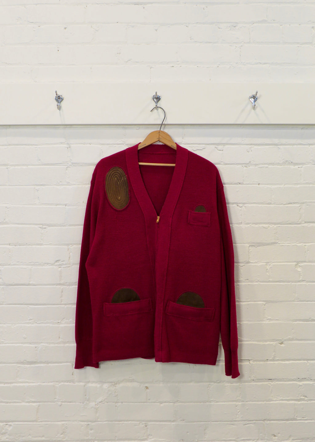Red cardigan with patches on a wooden hanger against a light wooden paneled wall.