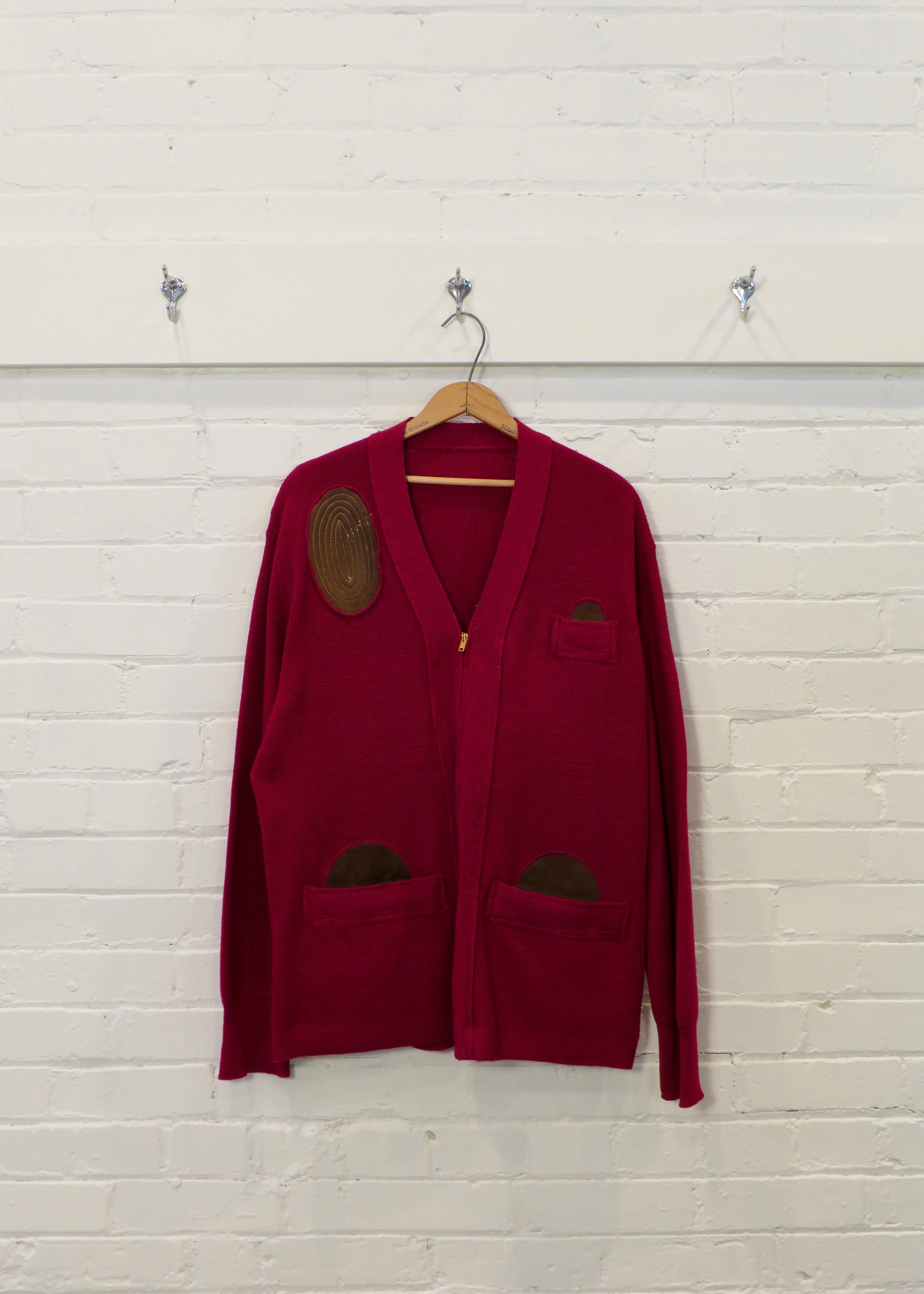 Red cardigan with patches on a wooden hanger against a light wooden paneled wall.